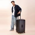 Man standing next to black Trunk Check-In Plus suitcase, stylish travel gear.