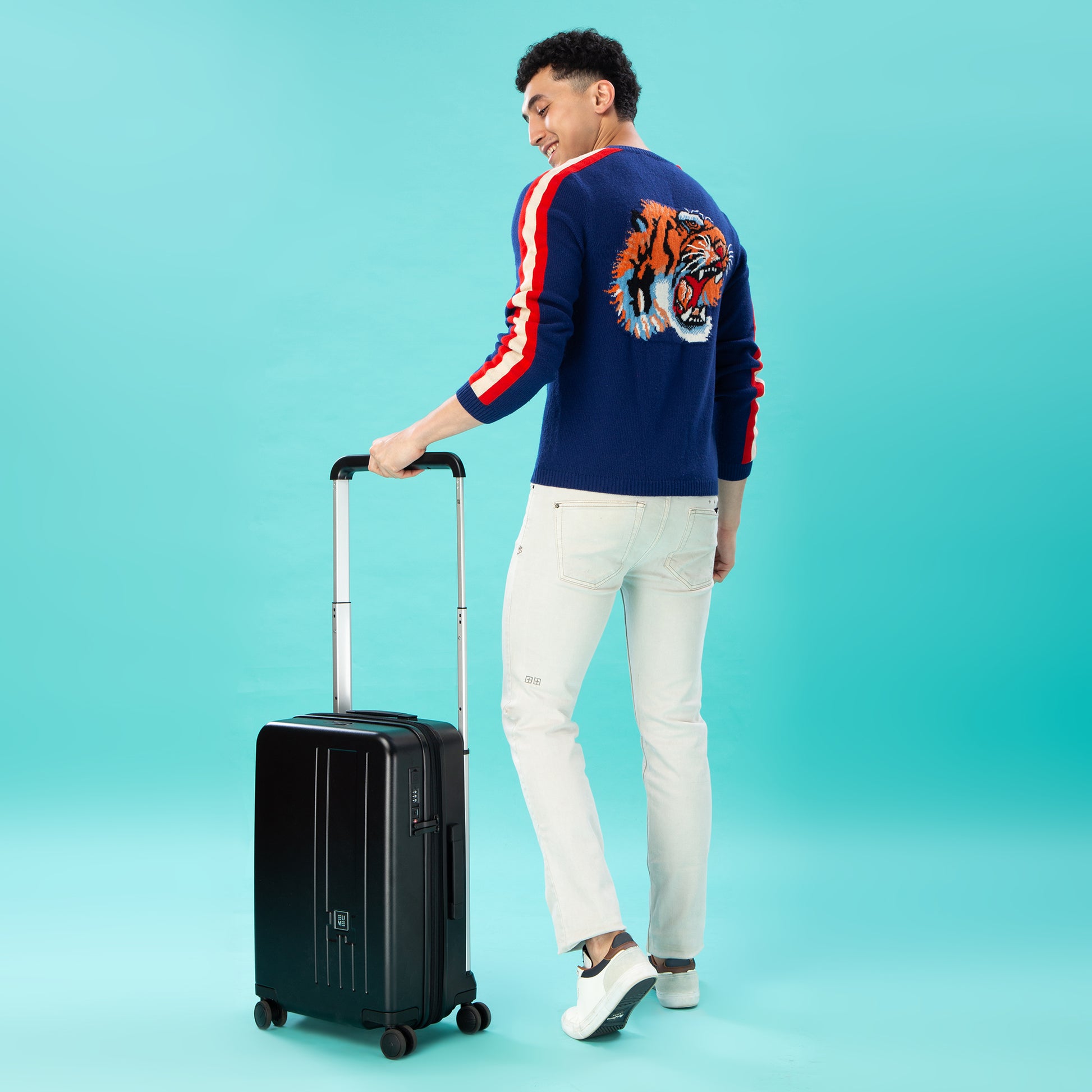 Man holding black suitcase while wearing stylish sweater against turquoise background.